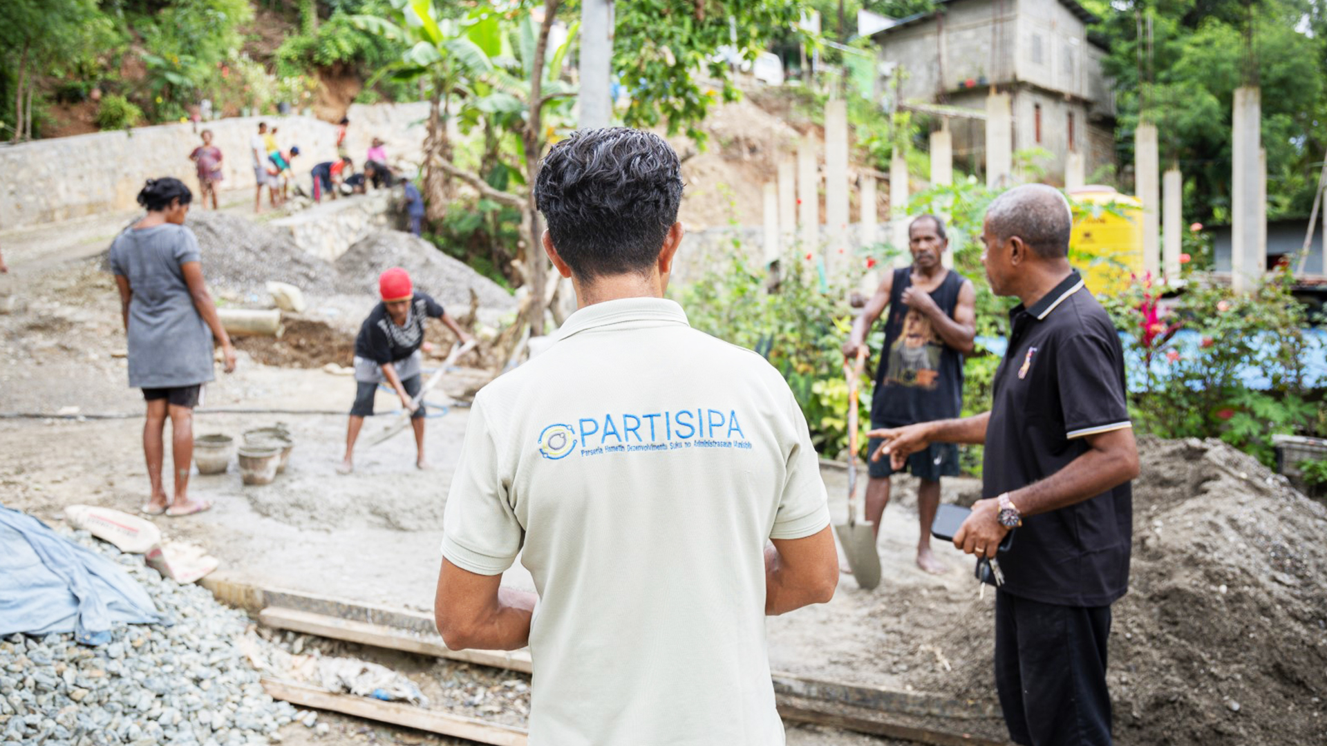 A staff member stands with his back to the camera, with PARTISIPA across the back of his t-shirt. He is surveying a group of community members as they work on a road that winds uphill. 