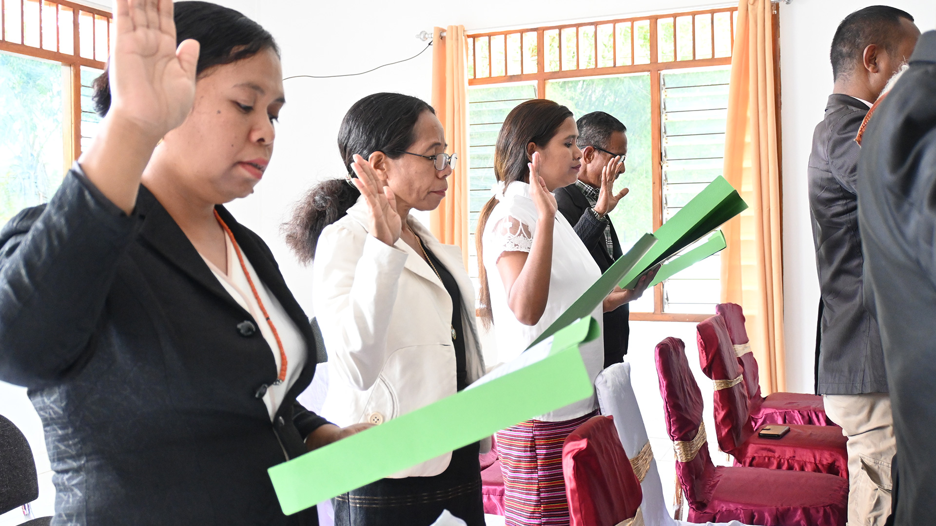A group of government workers are standing in a line with their right arm raised, holding green folders during a government meeting.