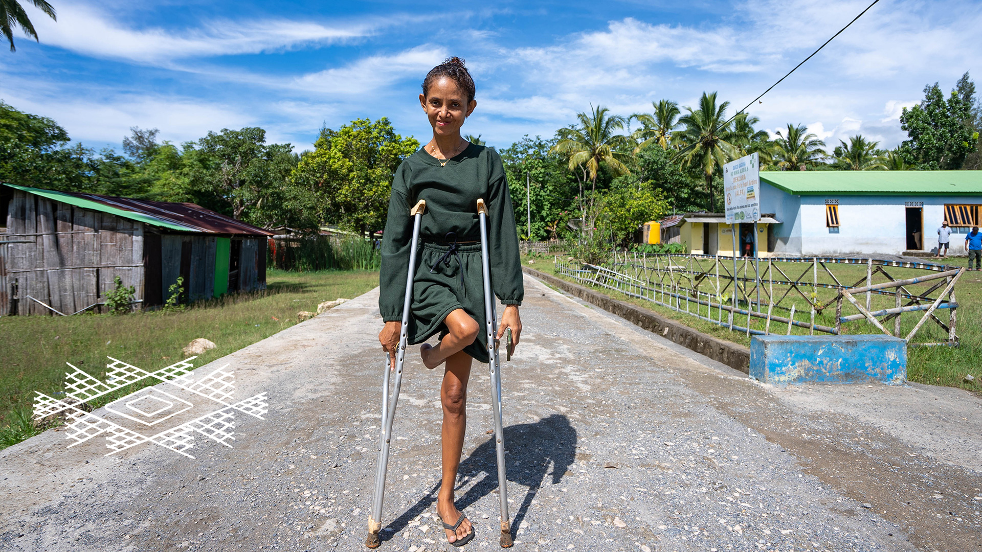 A smiling woman stands with crutches in the middle of a road in a Timorese village. There are buildings and people on the side of the road and trees in the background.