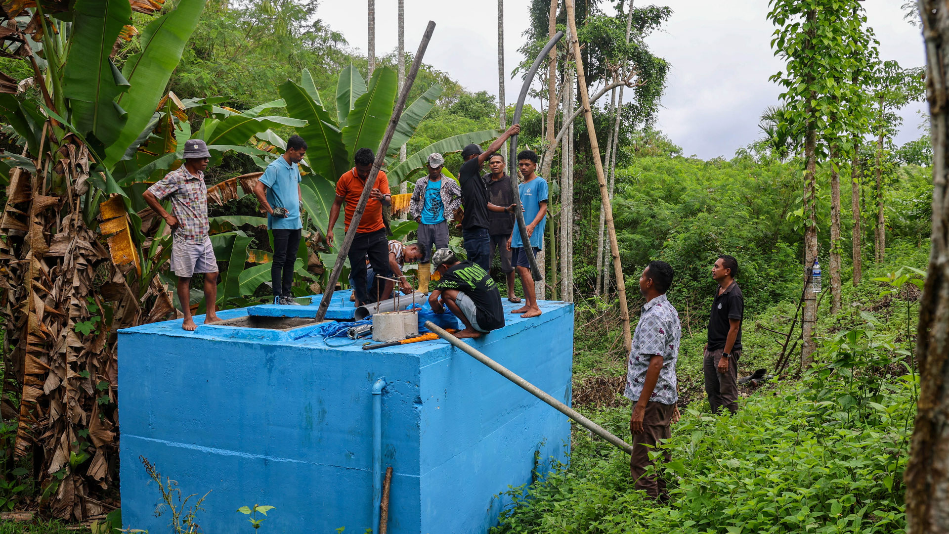 A group of villagers stand on top of a large concrete water tank painted blue and set amongst a lush tropical environment, they are conducting repairs.