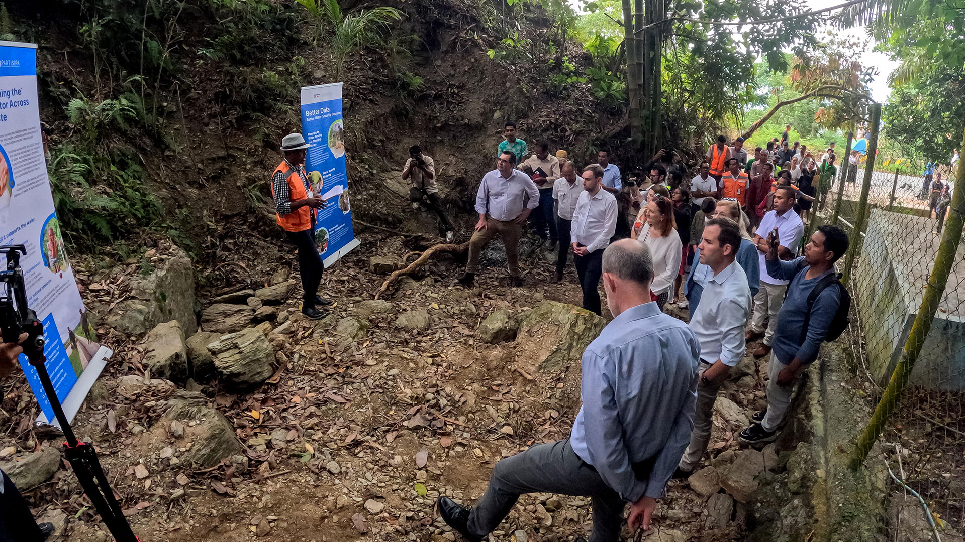 A group of Australian people long with PARTISIPA staff and communitiy members are standing spread up a hillside next to a water reservoir in Dare. A man in an orange high visiblity vest is presenting to them with two pull up banners displaying more information.