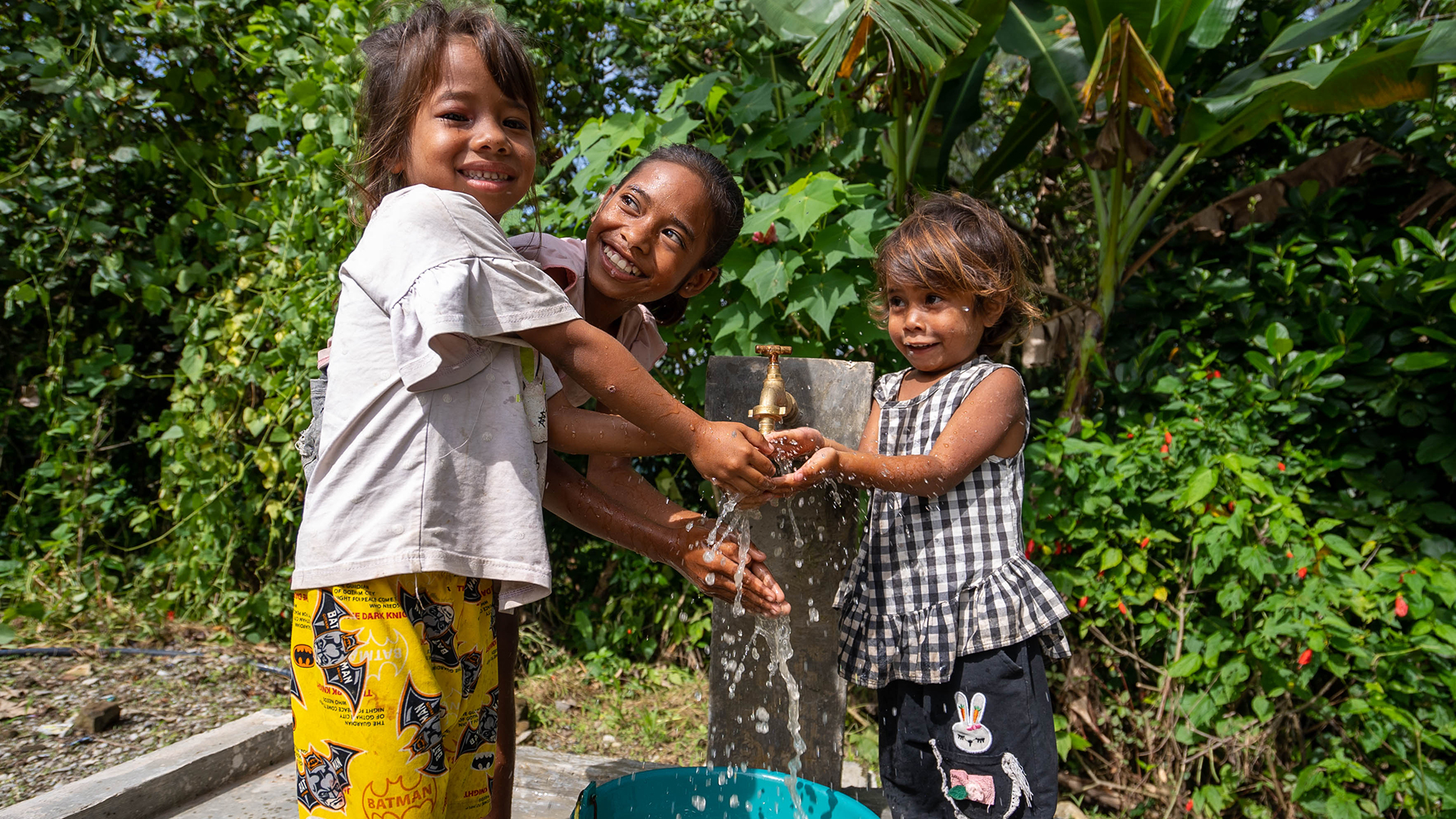 A group of three happy children smile as water gushes from a tap stand into their open hands. There are lush green plants in the background.